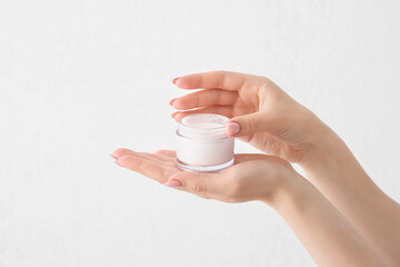 Hands of young woman with jar of cream against light background