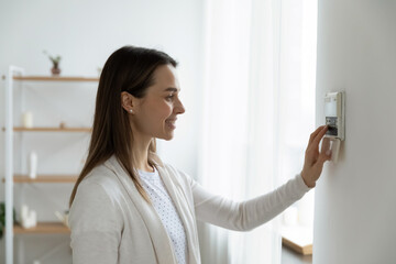 Smiling woman standing near wall-mounted device adjusting degrees in living room set comfortable...