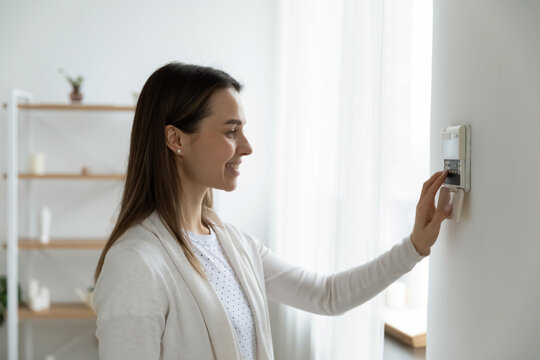 Smiling woman standing near wall-mounted device adjusting degrees in living room set comfortable temperature using thermostat home heating system. Owner of modern smart house, energy saving concept