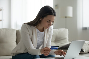 Serious woman sit on sofa typing on laptop holding plastic credit card using secure payment...