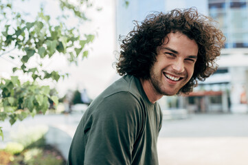 Cheerful young man with curly hair, smiling with healthy white teeths, sitting on the bench in the city street. Happy student hipster male has joyful expression while resting outside.