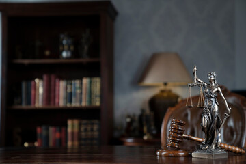 Judge chamber. Gavel and Themis statue on brown shining desk. Collection of legal books in the bookshelf.