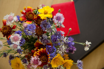 A bouquet of wildflowers is on the table against the background of a red notebook and laptop. View from above.
