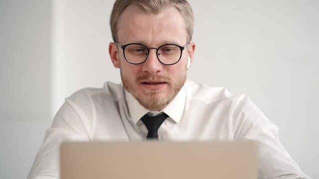 Serious young businessman look at laptop screen and talk on conference. calling by webcam, focused discuss during videoconference at home office. drink coffee cup. wireless headphone. ask answer