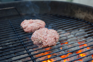 A view of two raw hamburger patties cooking on a kettle barbecue. Glowing embers and charcoal are...