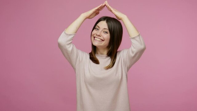 I'm in safety! Friendly charming brunette woman standing under hands roof gesture and smiling to camera, life insurance policy, wishing own home. indoor studio shot isolated on pink background