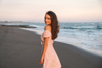 A beautiful young tourist girl walks along an empty black sand beach by the ocean in Bali. The girl in the dress smiles and turns around.
