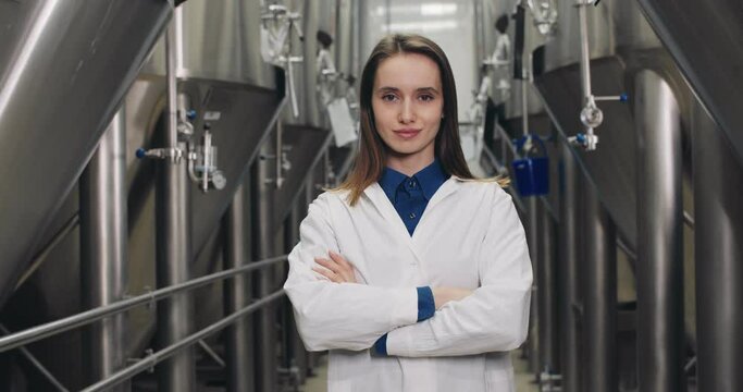 Crop view of attentive female worker looking to camera. Good looking woman in white lab coat standing at rows of steel brewing vats and crossing hands .Concept of brewery and beer plant