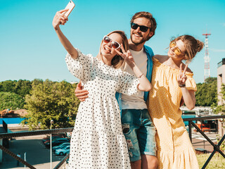 Group of young three stylish friends in the street.Man and two cute girls dressed in casual summer clothes.Smiling models having fun in sunglasses.Women and guy making photo selfie on smartphone