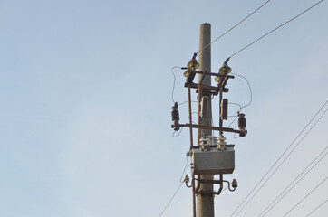 Old voltage transformer with safety and switching equipment on a metal frame on top of a concrete electric pole against a light blue sky in the open air.