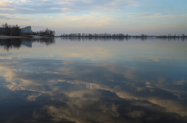 Landscape in the bay at the Dnieper river, Ukraine. Reflection of blue sky and light pink clouds in calm water. Buildings and trees on the shore on the horizon. Quiet March spring evening.