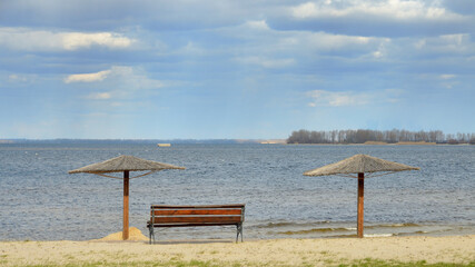 An empty bench and two reed umbrellas on the sandy shore of a pond against water in the spring in March. Blue cloudy sky. Forest on the horizon.