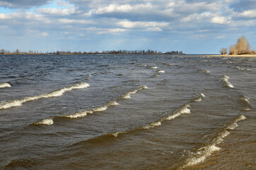 A series of running waves with white crests in shallow water in the foreground. Cloudy sky over the horizon. Landscape on the right bank of the Dnieper River in March, Ukraine.