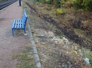 Bench at the polluted platform of a suburban railway station. Trash on the slope of the embankment...