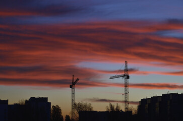 Two construction tower cranes at a background of scarlet dark clouds and blue sky. Silhouette of buildings and trees against a bright sky at dawn in November.