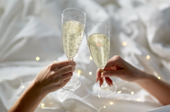 Lgbt, Same-sex Marriage, Celebration And Love Concept - Close Up Of Female Gay Couple Hands Holding And Clinking Champagne Glasses In Bed