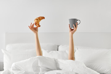 morning, comfort and people concept - young woman with cup of coffee and croissant lying in bed at...