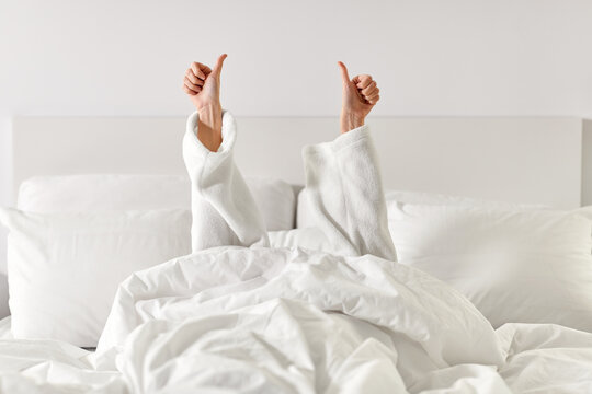 gesture, comfort and morning concept - hands of young woman in hotel robe lying in bed and showing thumbs up at bedroom
