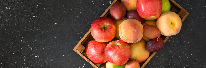 Various fruits in a wooden box on the black kitchen table. Apples, plums and peaches in a brown wooden box. Top view with space for text. Banner