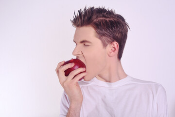 A man in a white shirt on a white background bites a red apple. The guy eats fruit and leads a healthy lifestyle