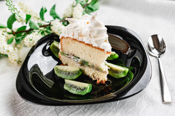 A slice of cake with meringue cream on a glossy black plate with slices of fresh kiwi. Cake decorated with a sprig of fragrant cherry blossoms