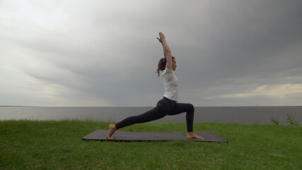 Fototapeta na wymiar Young fit woman practice yoga on coast near the lake or sea. Woman doing Warrior I Pose