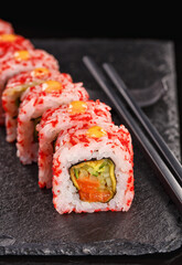 Uramaki sushi rolls covered in red caviar filled with salmon and cucumber. Served on a black stone board.