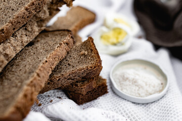 Slices of black bread and a boiled chicken egg on a white towel on the green grass. Picnic in nature with simple food. Next to the salt and flowers with plants in a wooden box.
