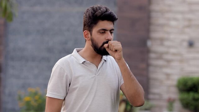 Portrait of an unhappy Indian guy coughing hard - common symptoms of Covid-19. A young attractive man coughs and keeps his fist near his mouth - Chinese coronavirus outbreaks in India