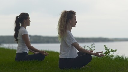 Two women meditating outdoors seated on the edge of coast near the sea, lotus pose folded fingers...