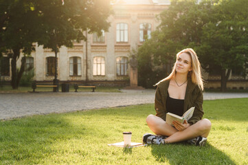 Cute pensive girl sitting on the grass in the university park with a book in hand ,looks away with...