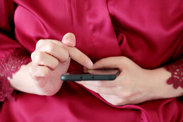 Woman using smartphone at home in a bed, girl in red lace nightie scrolling the screen of mobile phone by finger. Concept of online addiction, sms, social media