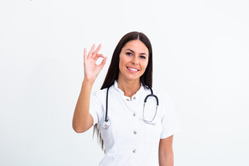 Medical concept of young beautiful female doctor in white coat with phonendoscope showing ok sign. Woman hospital worker looking at camera and smiling, studio, white background