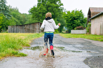 Happy kid boy wearing red rain boots and walking during sleet and rain on rainy cloudy day. Child in colorful casual clothes jumping into puddle. Having fun outdoors, healthy children activity