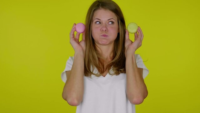 Funny woman in a white T-shirt looks at a delicious pink and yellow macaroons in hands with pouting cheeks, mouth on a yellow background with copy space. Place for text or product. Slowmotion
