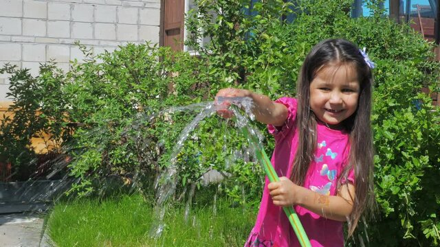 Little cute girl holds a watering hose in her hands. A child watering plants in the garden. The concept of childhood and motherhood