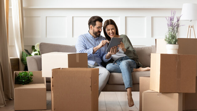 Excited young couple having fun with tablet on moving day, looking at mobile device screen, resting on couch in living room with cardboard boxes with belongings, relocation and mortgage concept