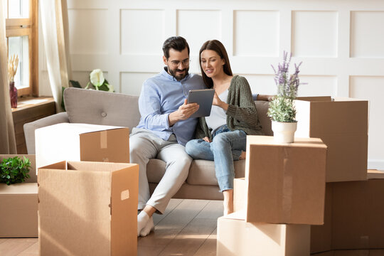 Smiling woman and man using computer tablet together, looking at screen, sitting on couch in living room with cardboard boxes with belongings, choosing moving service for relocation into new house