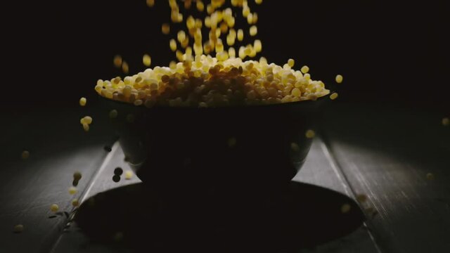 Grains of couscous falling into a black bowl on a table. Dark wooden surface. Slow motion.
