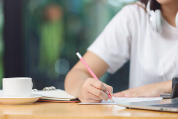 Asian woman student sitting at the table in living room, writing and using computer laptop when studying online course