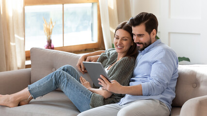 Happy young couple relaxing on cozy couch, using computer tablet together, smiling beautiful woman...