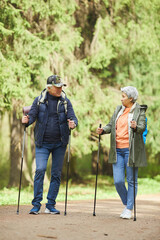 Fototapeta premium Full length portrait of active senior couple enjoying Nordic walking with poles during hike in forest