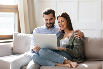 Happy young couple using laptop together, looking at screen, watching movie or video, shopping or...