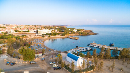 Fototapeta premium Aerial bird's eye view of coastline sunset, landmark white washed chapel Agia Triada beach, Protaras, Famagusta, Cyprus from above. Tourist attraction Ayia Trias bay church, sand, port at sunrise.