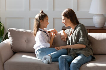Caring mother and little daughter chatting, sitting on cozy couch in living room, pretty girl...