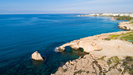 Fototapeta premium Aerial bird's eye view of coastline landmark ‘Love bridge’ and sea caves at Cavo Greco, Ayia Napa, Famagusta, Cyprus from above. A tourist attraction cliff rock natural formation arch in Ammochostos.