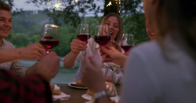 Group of happy friends celebrating holiday vacation using sprinklers and drinking red wine while having picnic french dinner party outdoor near the river