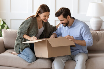 Happy curious couple unpacking parcel at home, sitting on couch, smiling woman looking into open...