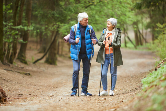 Full length portrait of active senior couple looking at each other and smiling while enjoying Nordic walk during hike in autumn forest, copy space - Powered by Adobe