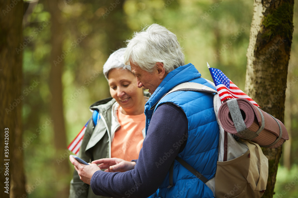 Wall mural side view at active senior couple using mobile app via smartphone while enjoying hike in forest, cop